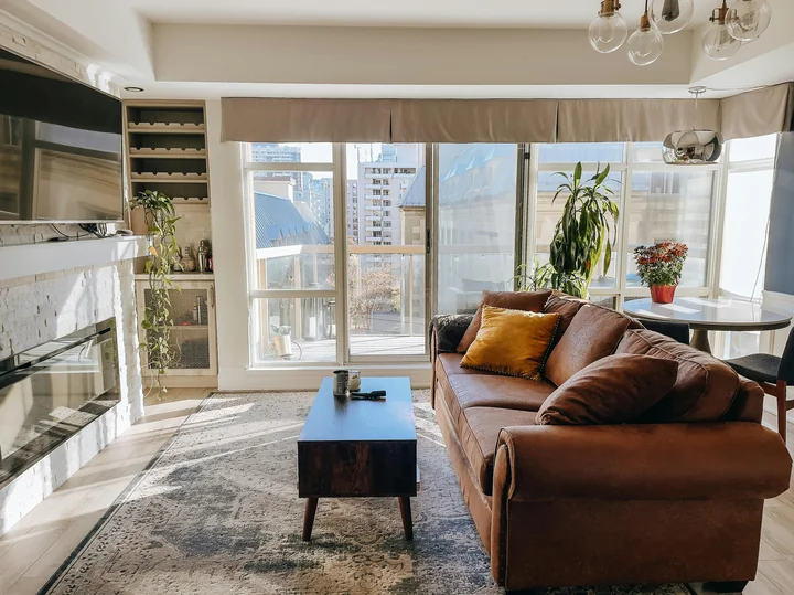 Jasne mieszkanie z dużymi oknami, brązową sofą z żółtą poduszką, stolikiem kawowym, roślinami i widokiem na miasto.