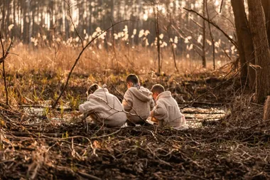 Troje dzieci w jasnych kurtkach siedzi przy strumieniu w lesie o zachodzie słońca.