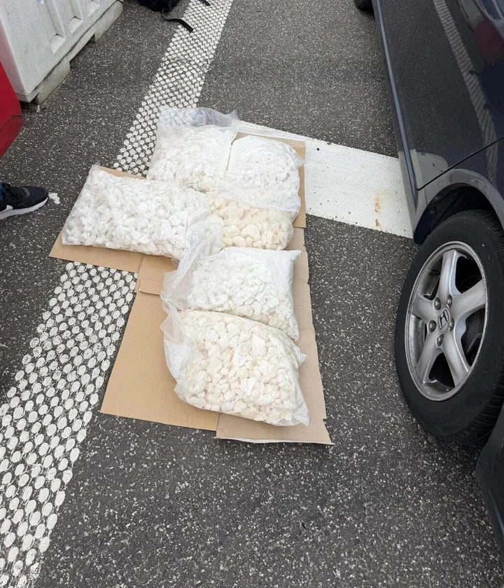 Woreczki z białymi tabletkami ułożone na kartonie na asfalcie obok samochodu osobowego.