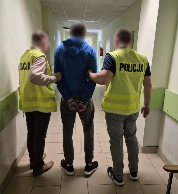 Dwaj policjanci w żółtych kamizelkach z napisem „POLICJA” prowadzą zatrzymanego mężczyznę w kajdankach przez korytarz budynku.