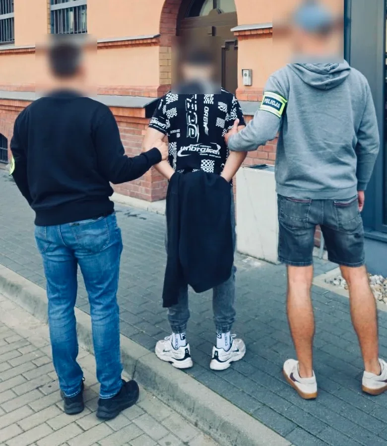 Funkcjonariusz z opaską POLICJA zatrzymuje mężczyznę na chodniku przed budynkiem z ceglaną elewacją, obok stoi kolejny mężczyzna.