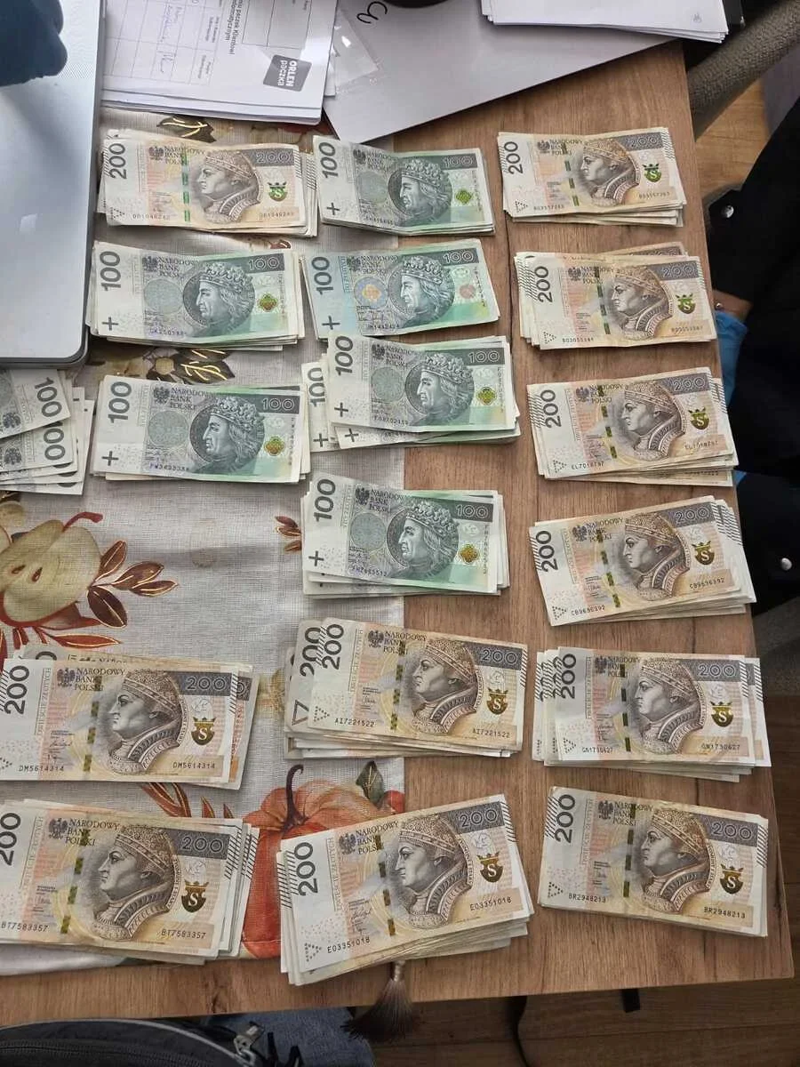Na drewnianym stole leżą ułożone w stosy banknoty o nominałach 100 i 200 złotych, obok dokumenty i laptop.