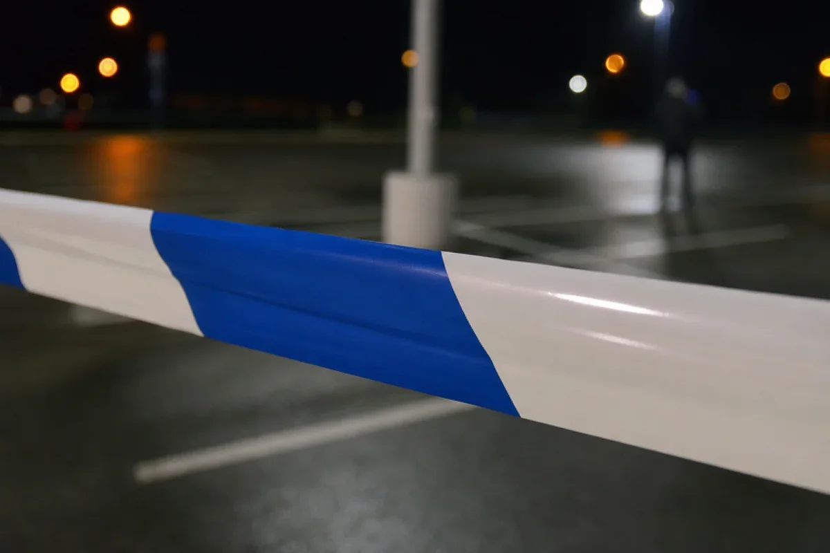 Nocna ulica z zamkniętą taśmą ostrzegawczą w biało-niebieskie pasy, w tle rozmyta postać przechodnia i światła uliczne.