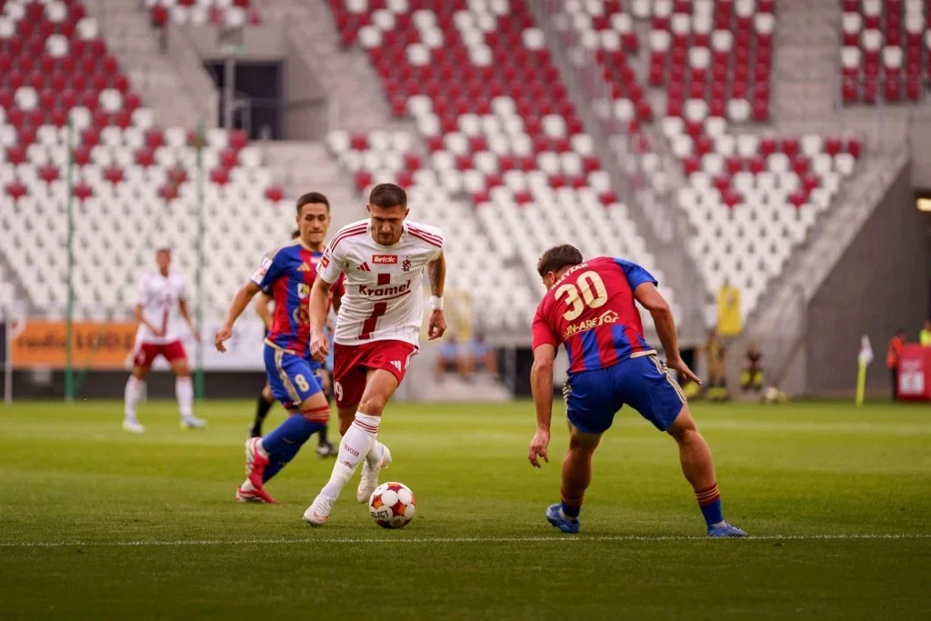 Piłkarz w białej koszulce z czerwonymi spodenkami prowadzi piłkę podczas meczu na stadionie, broniony przez dwóch rywali w niebiesko-czerwonych strojach.