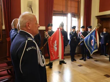 Funkcjonariusze Policji w galowych mundurach uczestniczą w uroczystości z sztandarami w pomieszczeniu o drewnianej boazerii.