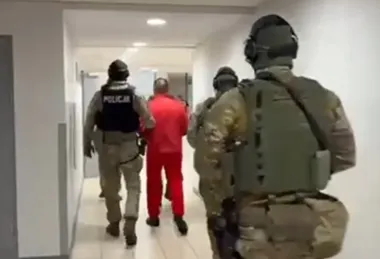 Funkcjonariusze policji w mundurach taktycznych prowadzą mężczyznę w czerwonym dresie przez korytarz budynku.