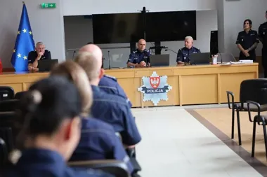 Policjanci w mundurach uczestniczą w naradzie lub odprawie w sali konferencyjnej z logo Policji na pulpicie.