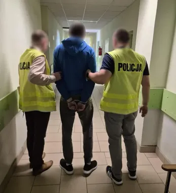 Dwaj policjanci w żółtych kamizelkach z napisem „POLICJA” prowadzą zatrzymanego mężczyznę w kajdankach przez korytarz budynku.