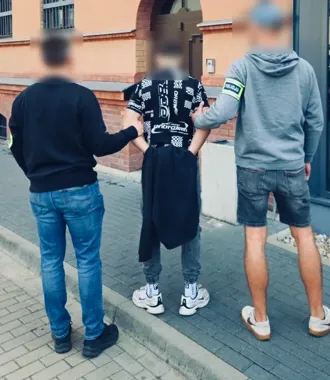 Funkcjonariusz z opaską POLICJA zatrzymuje mężczyznę na chodniku przed budynkiem z ceglaną elewacją, obok stoi kolejny mężczyzna.