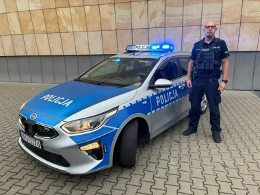 Funkcjonariusz policji w mundurze stoi obok oznakowanego radiowozu Kia z włączonymi niebieskimi światłami, zaparkowanego przed budynkiem z płytkami elewacyjnymi
