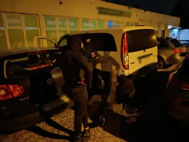 W nocy na parkingu przed budynkiem osoba w kapturze stoi nad dwiema osobami siedzącymi na ziemi przy białym samochodzie dostawczym.