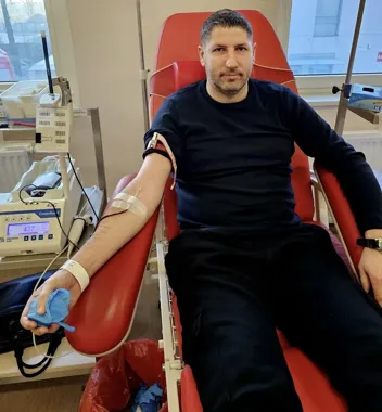 Mężczyzna siedzi na czerwonym fotelu w punkcie krwiodawstwa z igłą w przedramieniu podczas pobierania krwi.