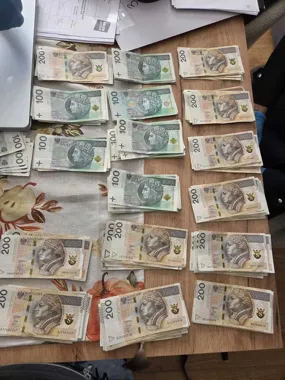Na drewnianym stole leżą ułożone w stosy banknoty o nominałach 100 i 200 złotych, obok dokumenty i laptop.