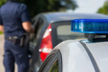 Policjant w mundurze stoi przy tylnej części ciemnego samochodu osobowego, na pierwszym planie widać policyjny radiowóz z niebieskim światłem.
