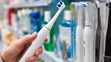 Ręka trzymająca białą elektryczną szczoteczkę do zębów na tle półek sklepowych z produktami higienicznymi.