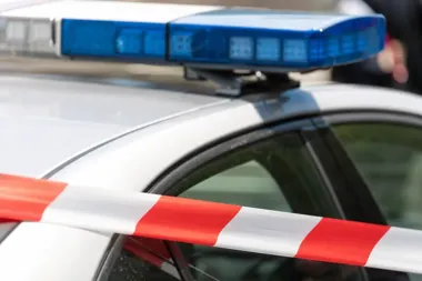 Fragment policyjnego radiowozu z niebieskim światłem na dachu, przed samochodem przewinięta jest czerwono-biała taśma ostrzegawcza.