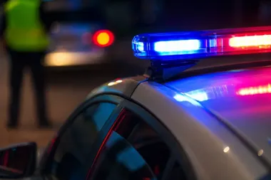 Nocna akcja służb: w pierwszym planie policyjny radiowóz z włączonymi niebiesko-czerwonymi światłami błyskowymi, w tle rozmyta postać funkcjonariusza w kamizelc