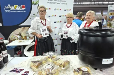 Trzy kobiety w regionalnych strojach stoją za stoiskiem z wypiekami podczas targów promujących województwo łódzkie.