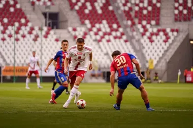 Piłkarz w białej koszulce z czerwonymi spodenkami prowadzi piłkę podczas meczu na stadionie, broniony przez dwóch rywali w niebiesko-czerwonych strojach.