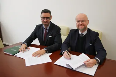 Dwaj mężczyźni w garniturach siedzą przy drewnianym stole i podpisują dokumenty w pomieszczeniu biurowym.