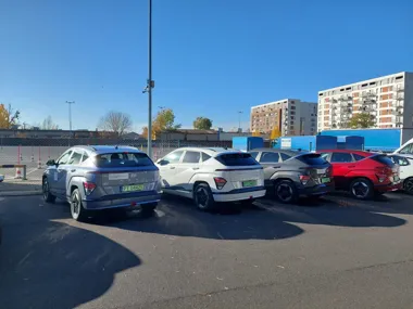 Cztery nowe samochody Hyundai Kona Electric w różnych kolorach stoją na parkingu przed budynkami mieszkalnymi w słoneczny dzień.